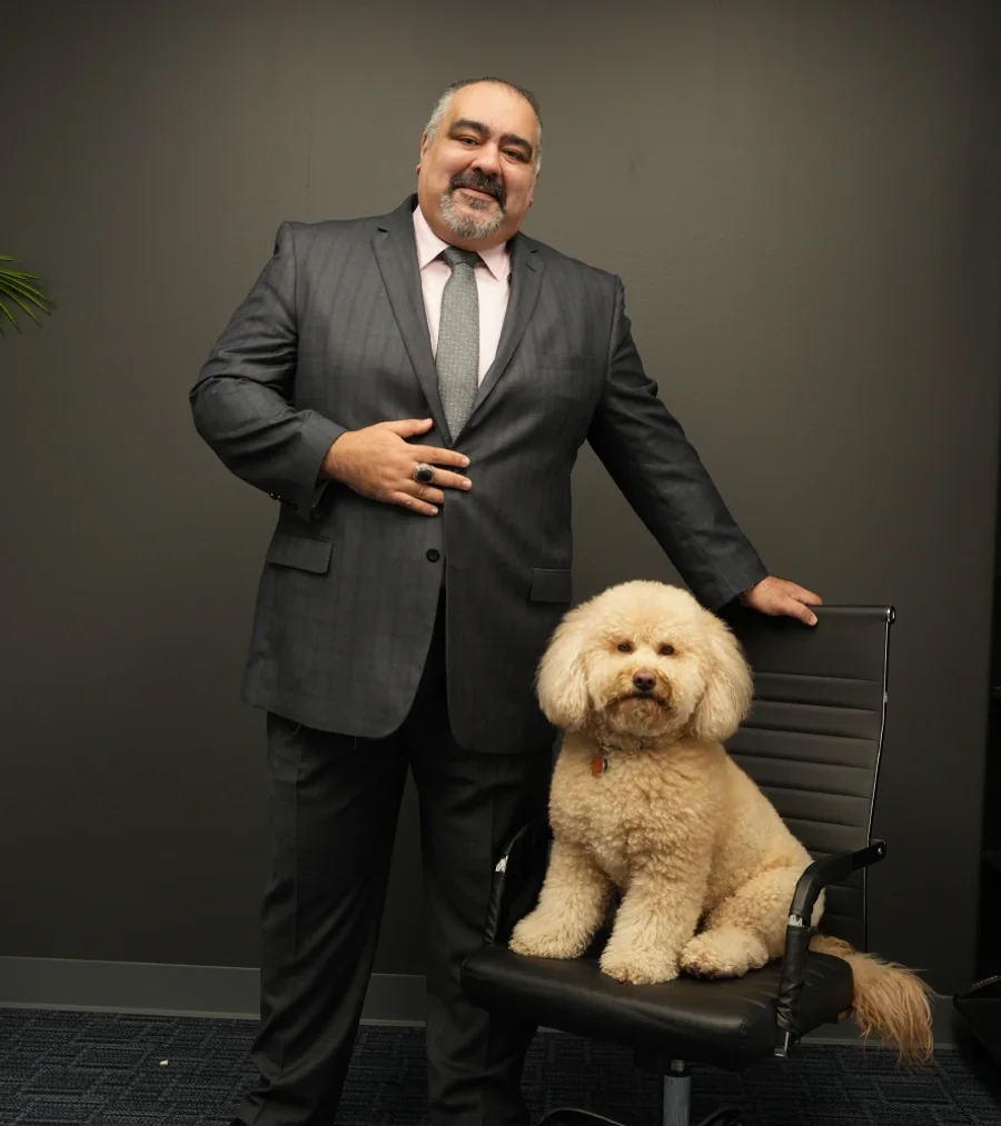Eman Afshar in a grey suit, standing beside his office chair with his dog sitting in it
